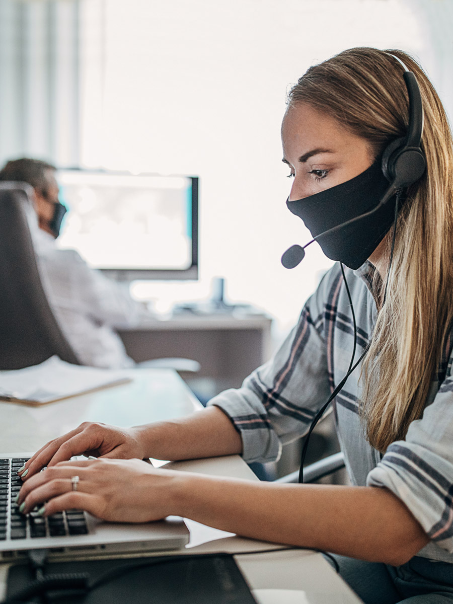 Frau sitzt mit Headset und Mund-Nasen-Schutz vor einem Computer